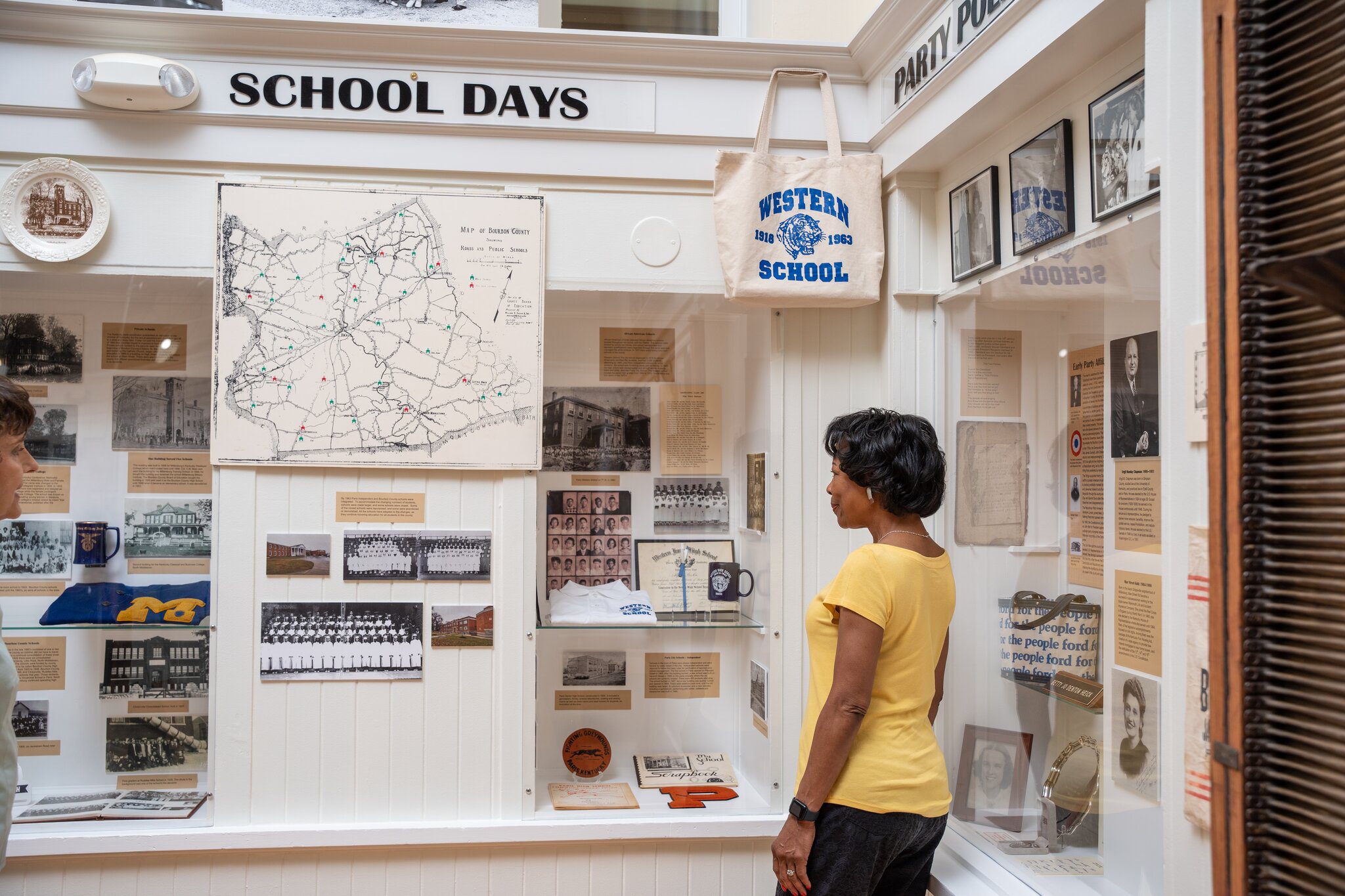 A woman in a yellow shirt looks at a School Days exhibit displaying vintage photos, documents, and memorabilia about local schools, including a map, trophies, and a tote bag reading Western School 1932.