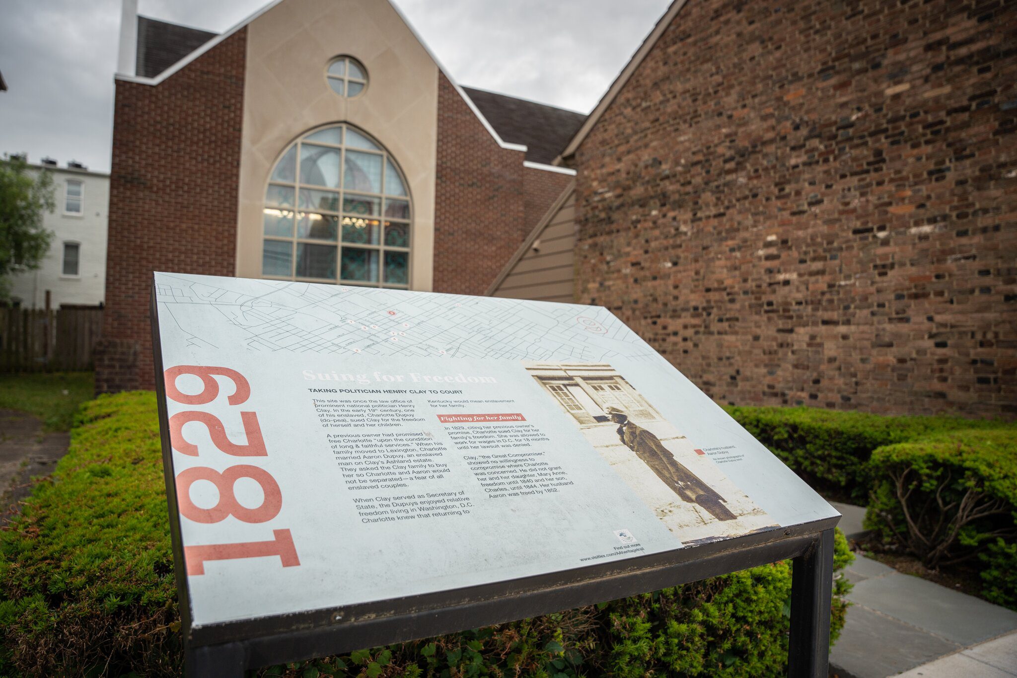 Informational sign titled 1829 stands in front of a brick building with large windows. The sign, surrounded by bushes, discusses the history of enslaved people seeking freedom.