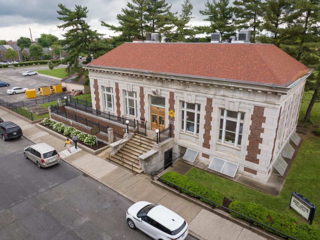 Aerial view of a rectangular brick and stone building with a red roof, large windows, and a central staircase entrance; cars are parked on the street and a person walks on the sidewalk.