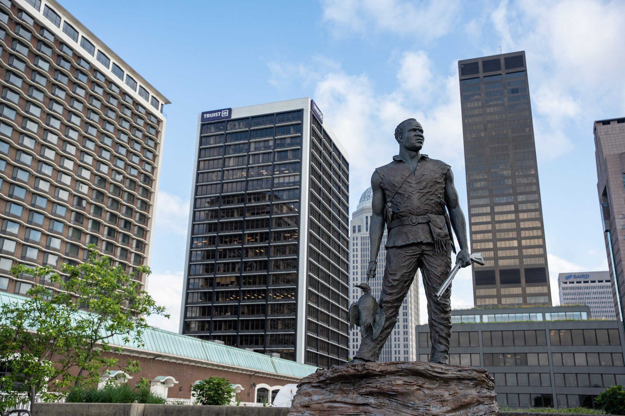 A bronze statue of a man standing on a rock with a dog by his side, set against a backdrop of modern tall office buildings in a cityscape under a partly cloudy sky.