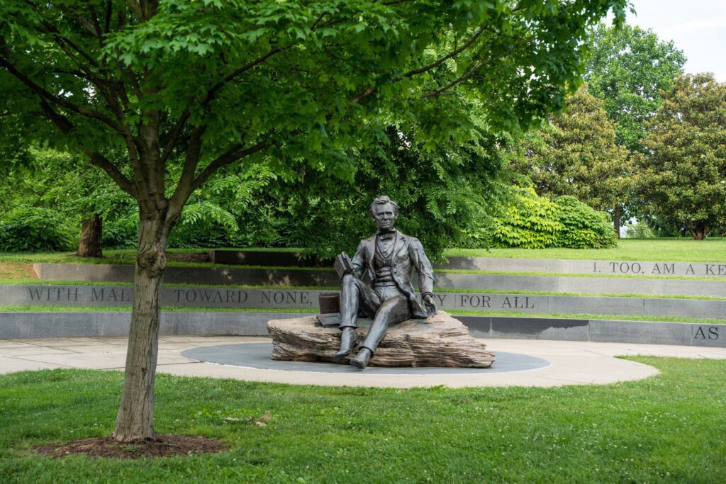 A bronze statue of Abraham Lincoln sits on a stone bench surrounded by greenery, with a curved wall behind it displaying engraved text. A leafy tree stands in the foreground.