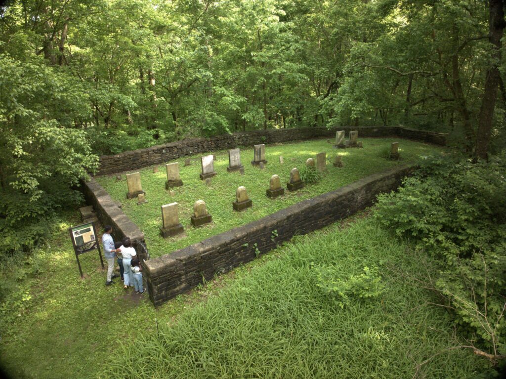 Four people stand outside a stone-walled, rectangular cemetery with old gravestones, surrounded by dense green trees and grass. The cemetery is in a forested area.