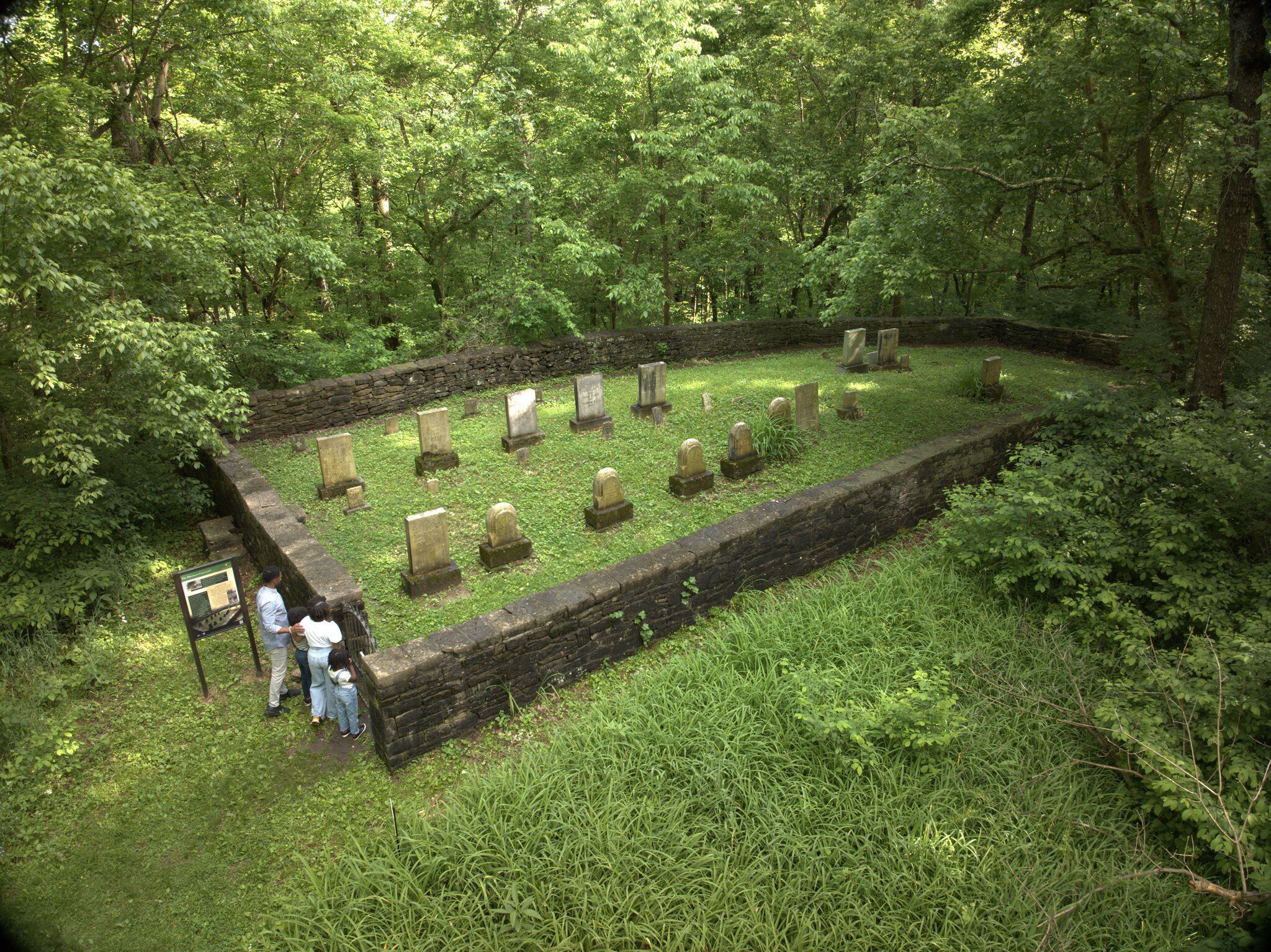 Four people stand outside a stone-walled, rectangular cemetery with old gravestones, surrounded by dense green trees and grass. The cemetery is in a forested area.