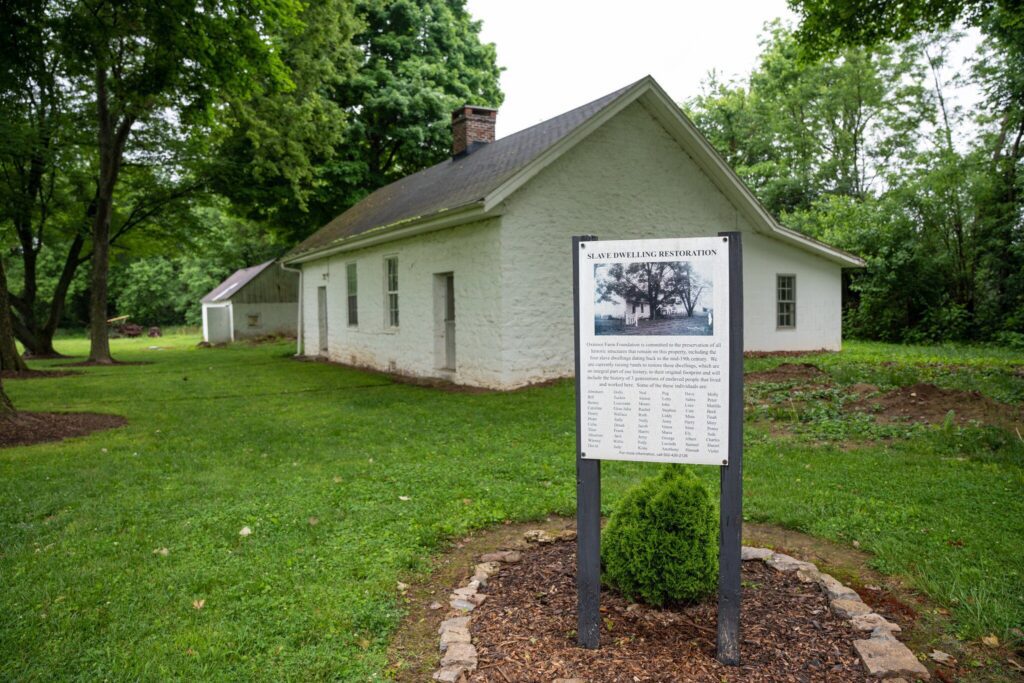 A historic white stone house stands surrounded by green grass and trees. In the foreground, a sign with information about the buildings restoration is displayed, bordered by a small circular garden.
