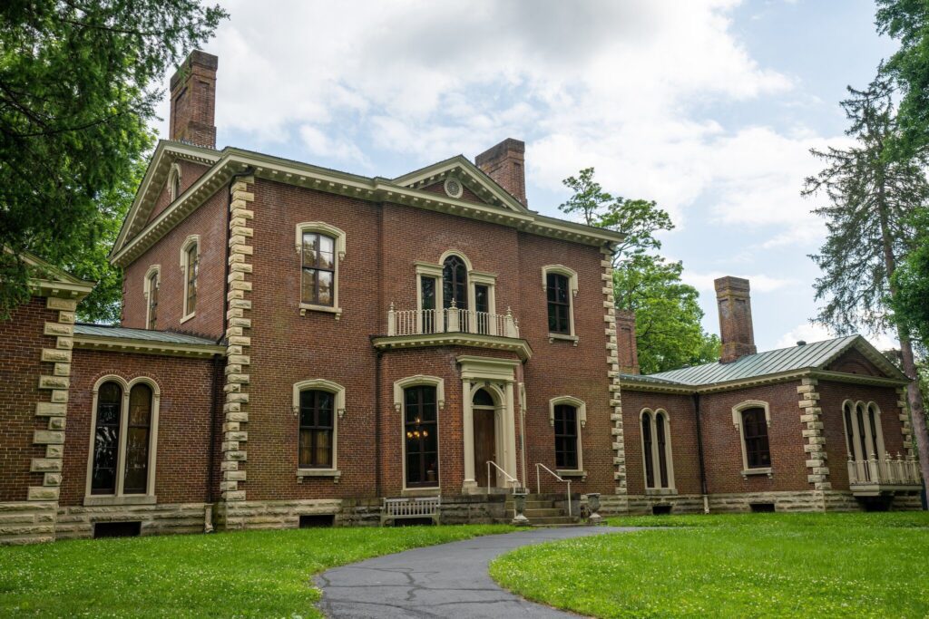 A two-story historic brick mansion with stone trim, tall windows, and a small central balcony, surrounded by trees and a lawn under a partly cloudy sky.