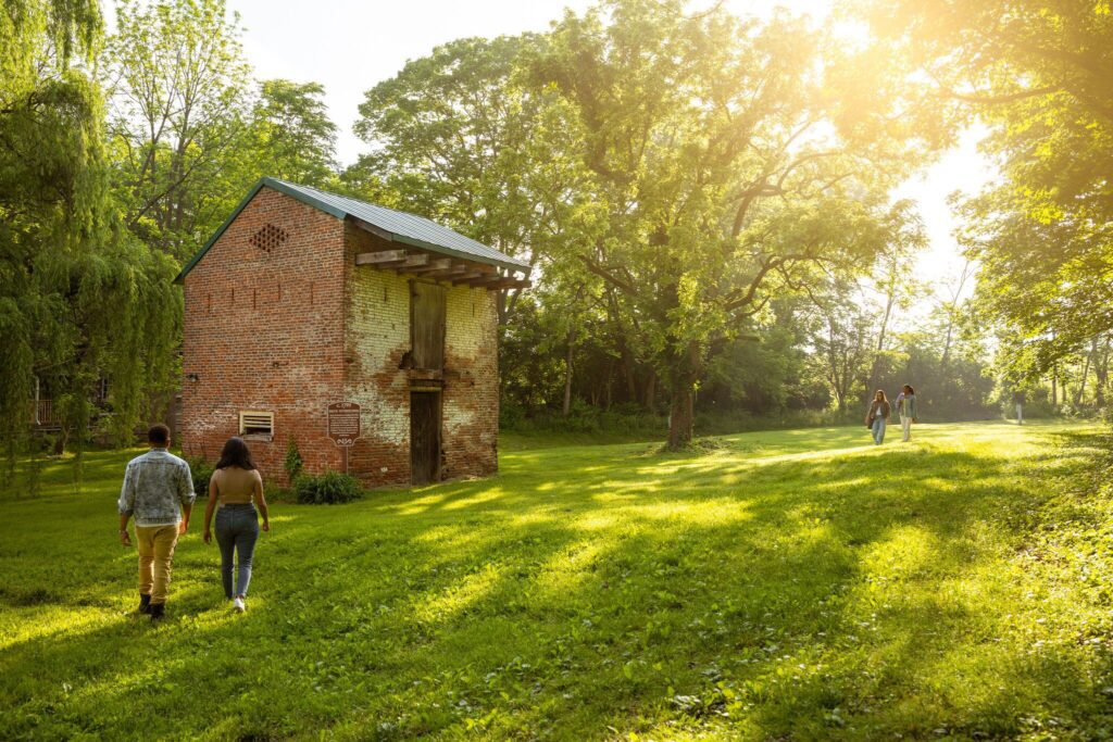 Two pairs of people walk across a sunlit grassy field toward and past a small old brick building, surrounded by tall trees and lush greenery on a bright, sunny day.