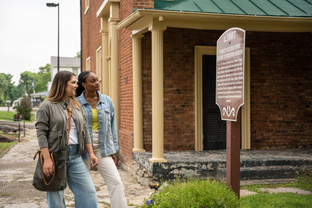 Two women stand outside a historic brick building, reading an informational sign. One holds a bag, and both appear interested and engaged with the sign. The setting is outdoors with greenery in the background.