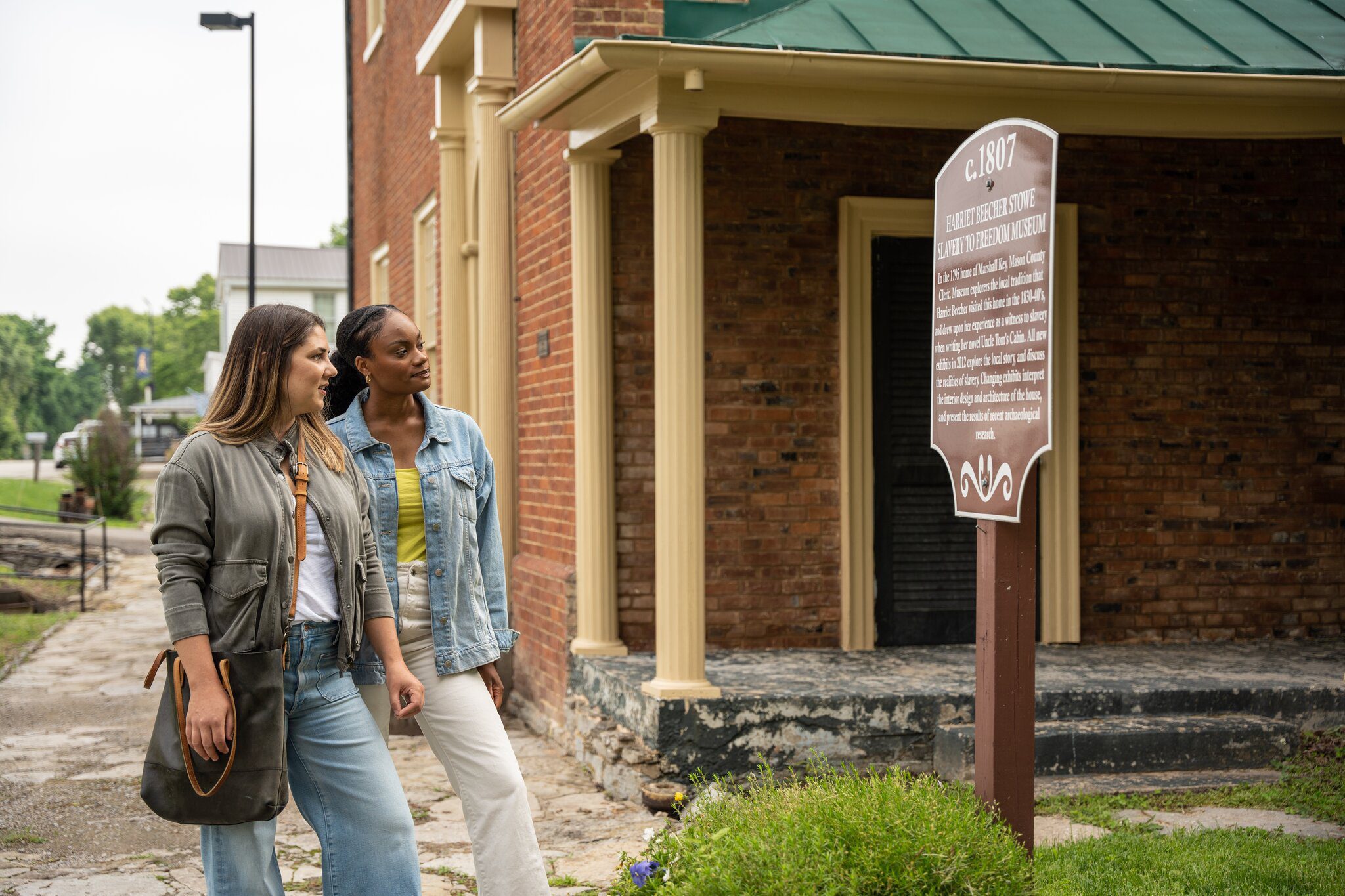 Two women stand outside a historic brick building, reading an informational sign. One holds a bag, and both appear interested and engaged with the sign. The setting is outdoors with greenery in the background.