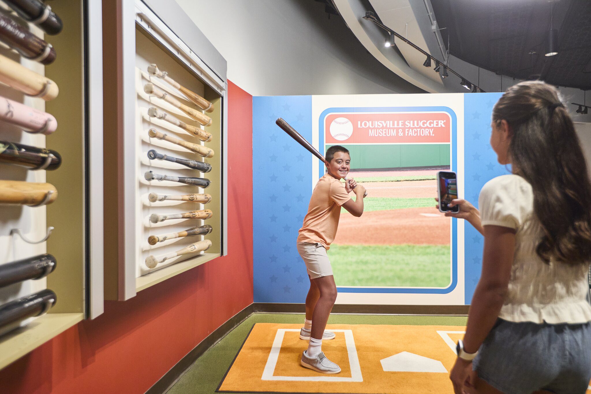 A person poses with a baseball bat in a batting stance at the Louisville Slugger Museum while another person takes their photo with a phone. Bats are displayed on the wall beside them.