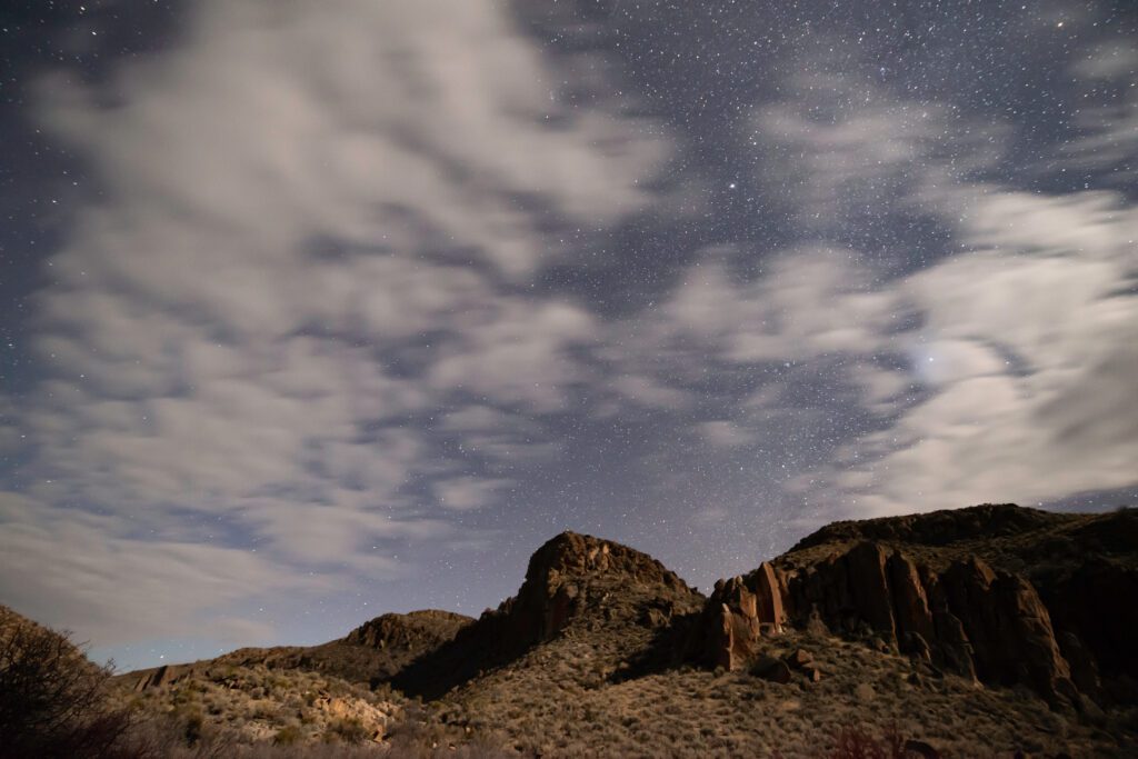 Clouds partially cover a star-filled night sky above rocky hills and desert shrubs, creating a dramatic and serene nighttime landscape.