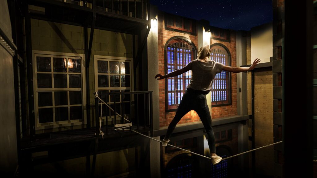 A person balances carefully on a tightrope above a dark, reflective surface indoors, with brick walls and windows lit by warm lights in the background.