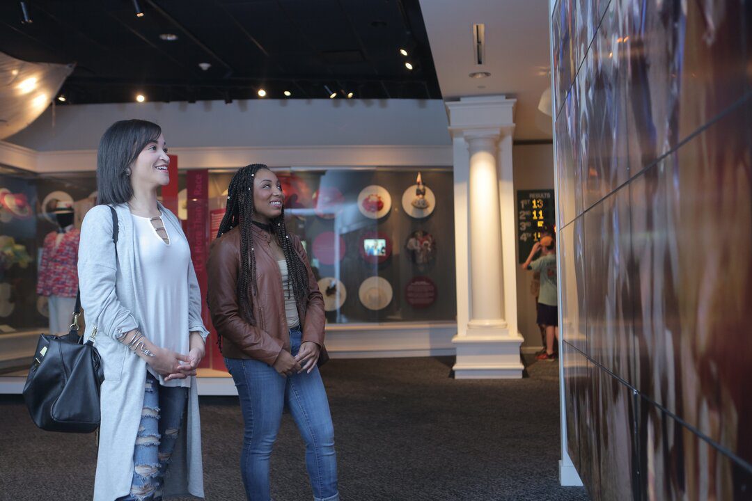 Two women stand side by side, smiling and looking at a large wall display in a museum exhibit. Displays with various objects are visible in the background.