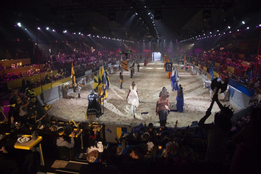 Performers in medieval costumes with flags stand in a sandy arena, surrounded by cheering spectators at themed tables, under dramatic lighting, recreating a medieval tournament scene indoors.