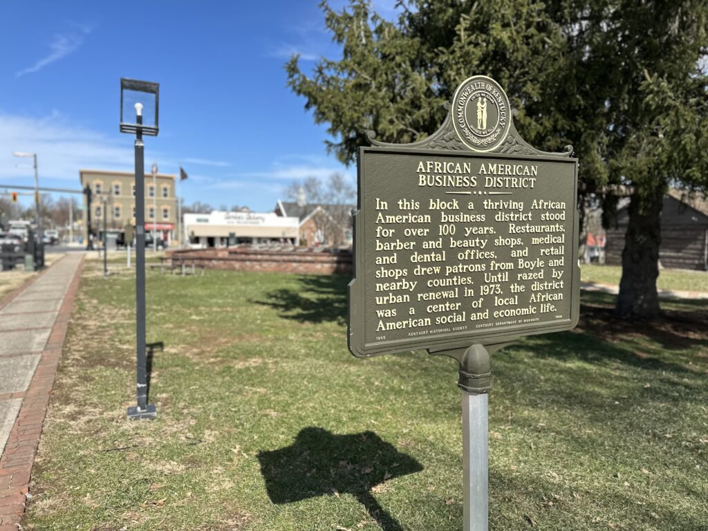 A historical marker reads “African American Business District,” describing the area’s significance. In the background are buildings, a sidewalk, trees, and a clear blue sky.
