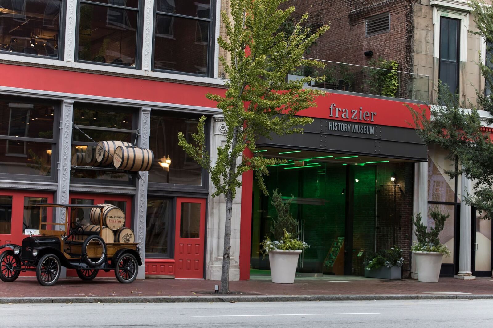Street view of the Frazier History Museum entrance with a vintage car carrying whiskey barrels parked outside. The building has large windows and red accents, with trees and plants along the sidewalk.