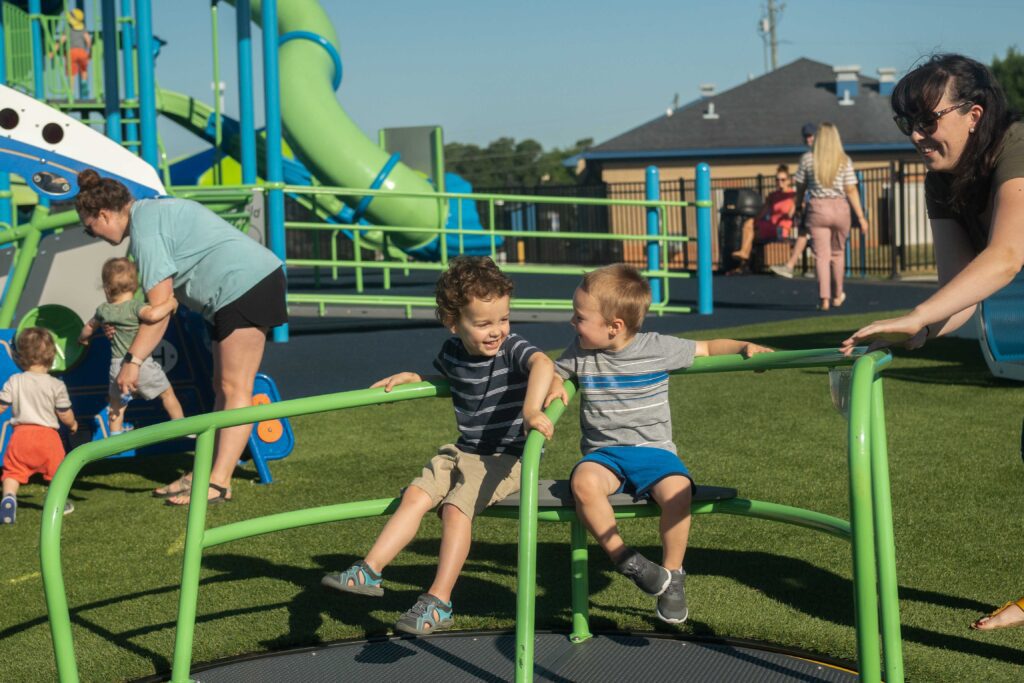 Two young boys laughing and sitting on a green merry-go-round at a playground. Adults and children are in the background near play structures and slides. The scene is sunny, with a bright blue sky.