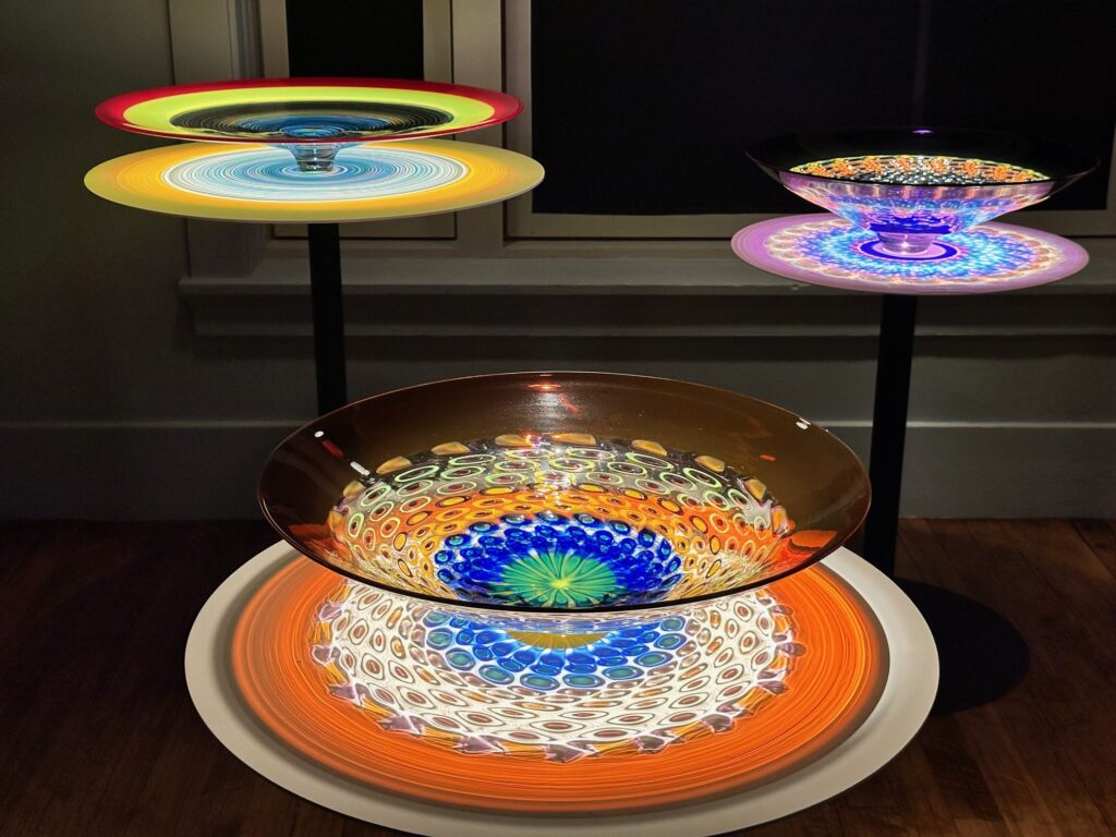 A display of four colorful, illuminated kaleidoscope-like discs mounted on stands. Each disc features intricate, symmetrical patterns in vibrant hues, with the central one having a bold blue core surrounded by concentric layers of yellow, orange, and white.