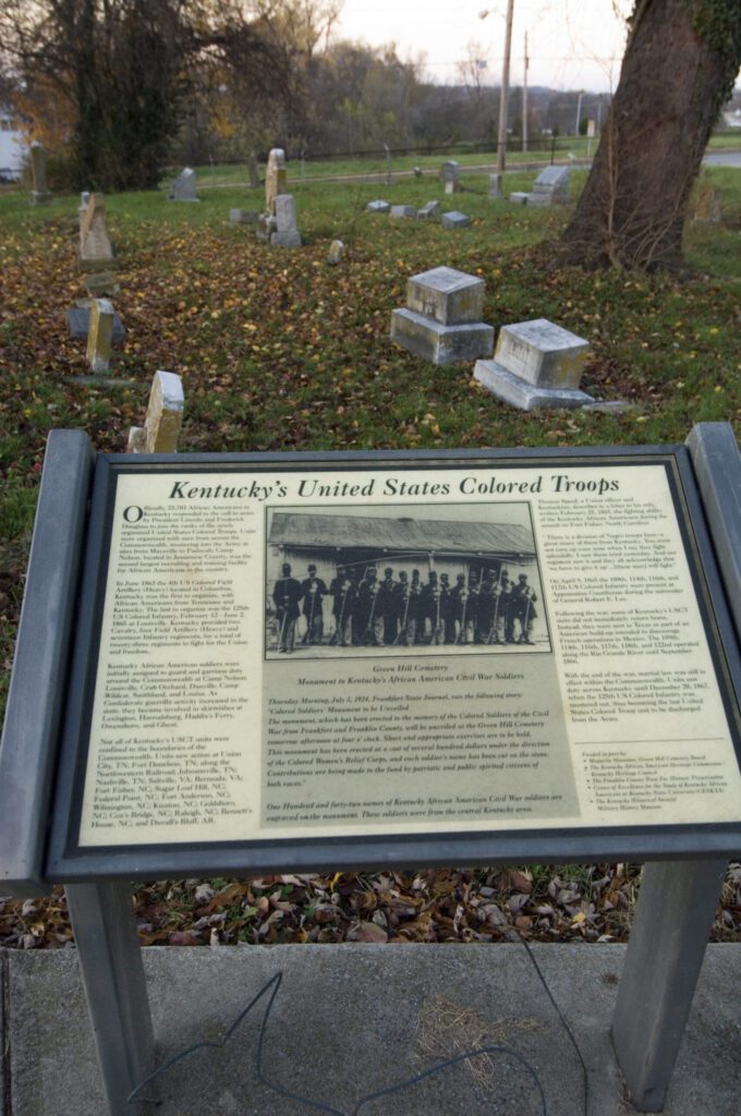 A plaque titled “Kentucky’s United States Colored Troops” stands in front of a cemetery with gravestones and fallen leaves scattered on the ground in the background.