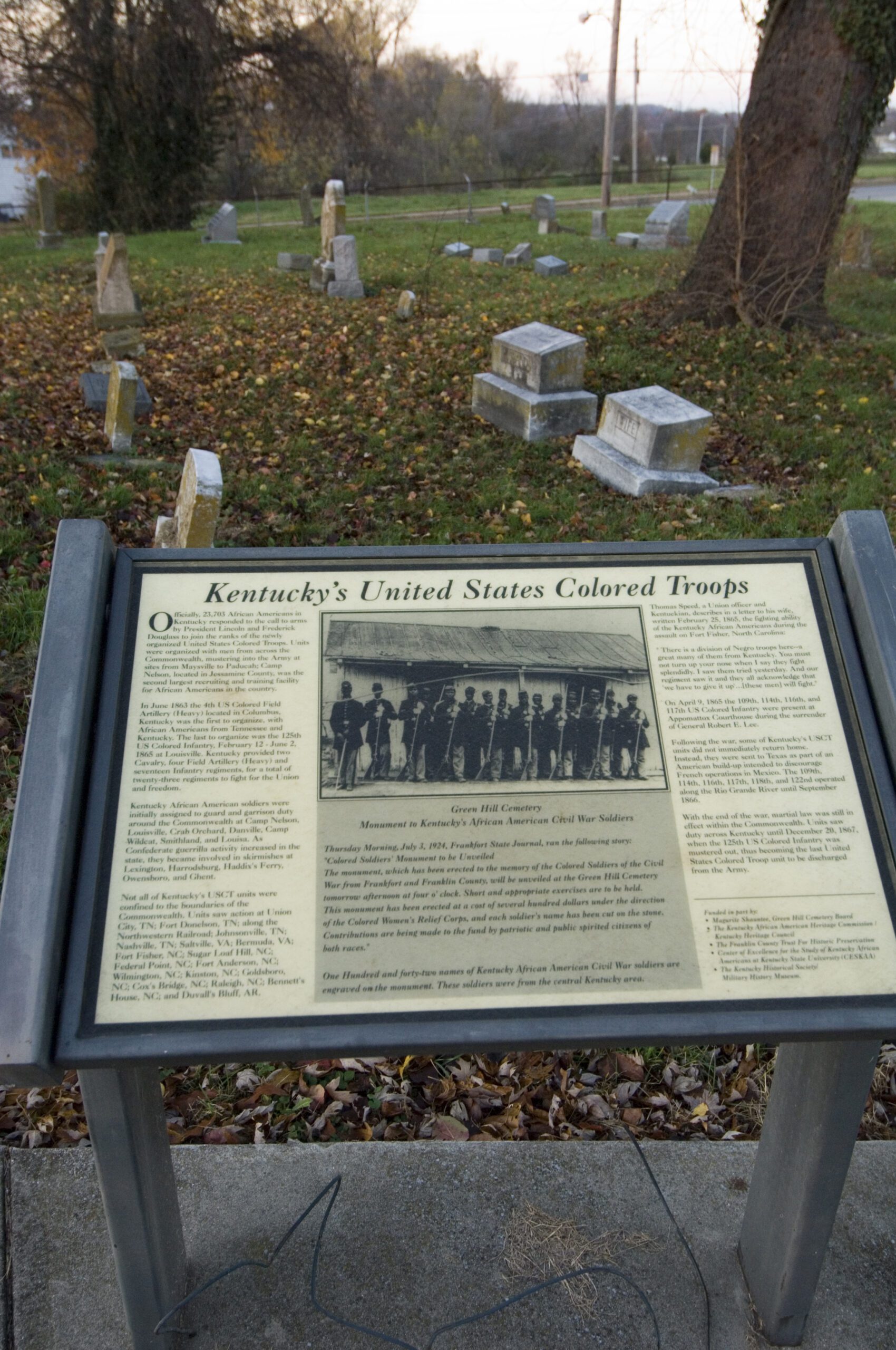 A plaque titled “Kentucky’s United States Colored Troops” stands in front of a cemetery with gravestones and fallen leaves scattered on the ground in the background.