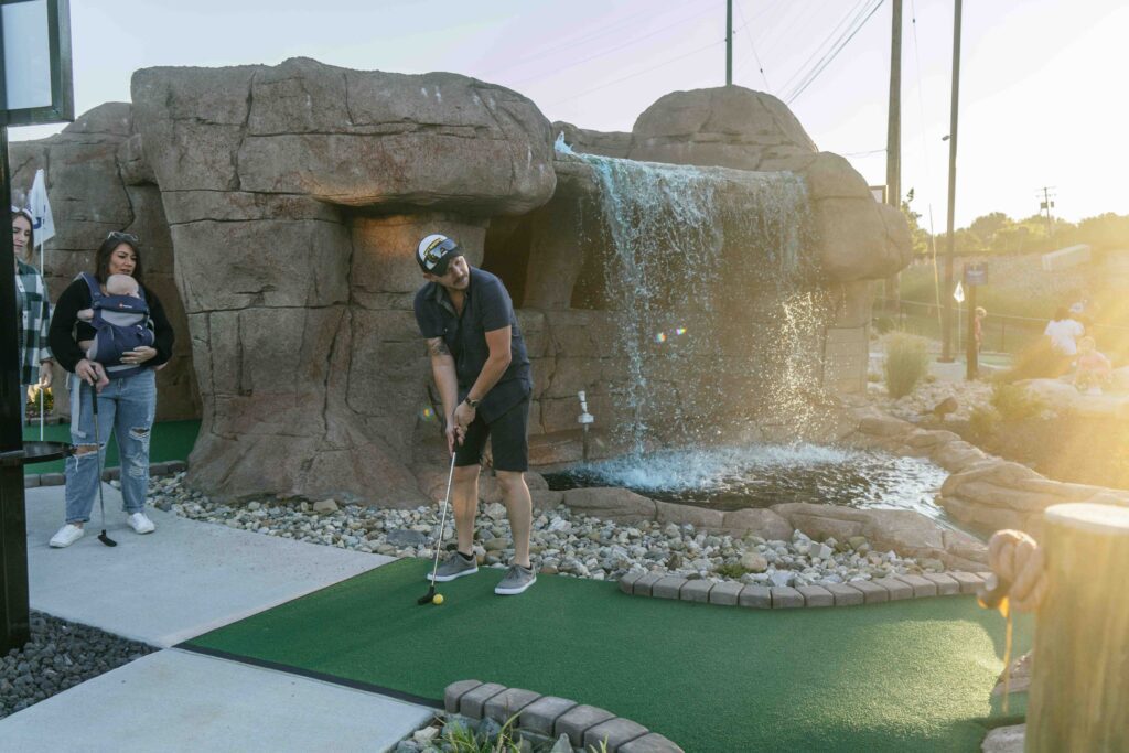 A person plays mini-golf on a course with a rock waterfall in the background. Another person holding a baby watches nearby. The setting sun casts warm light over the scene.