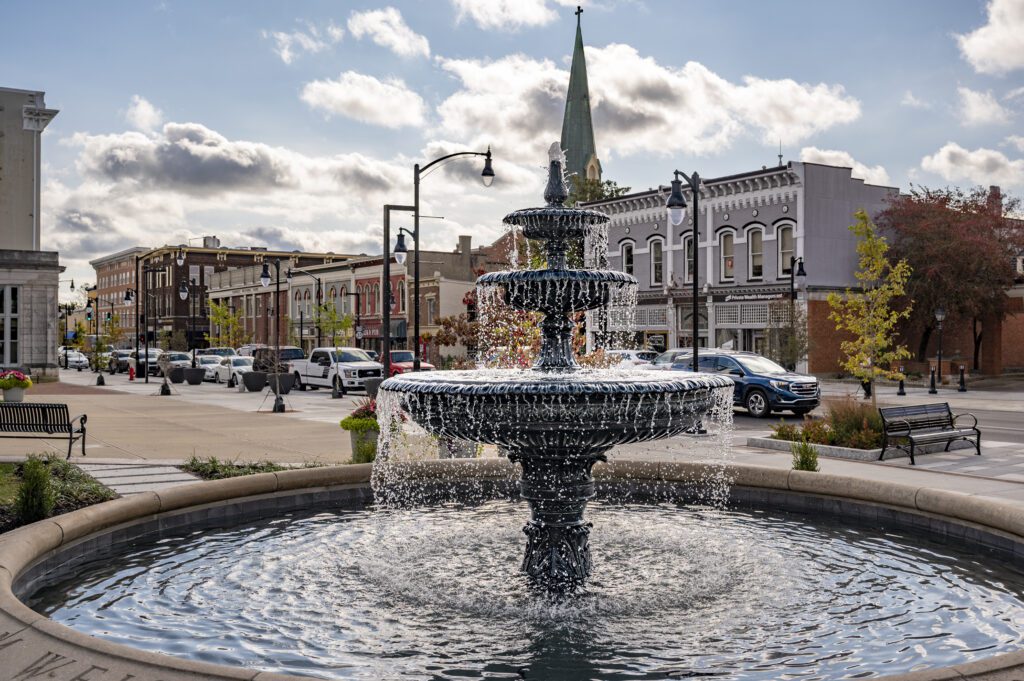 A tiered fountain in a round basin is in the foreground, with water cascading down. Behind it, there’s a street lined with brick buildings, trees with fall foliage, and cars. A church steeple rises against a sky filled with clouds.