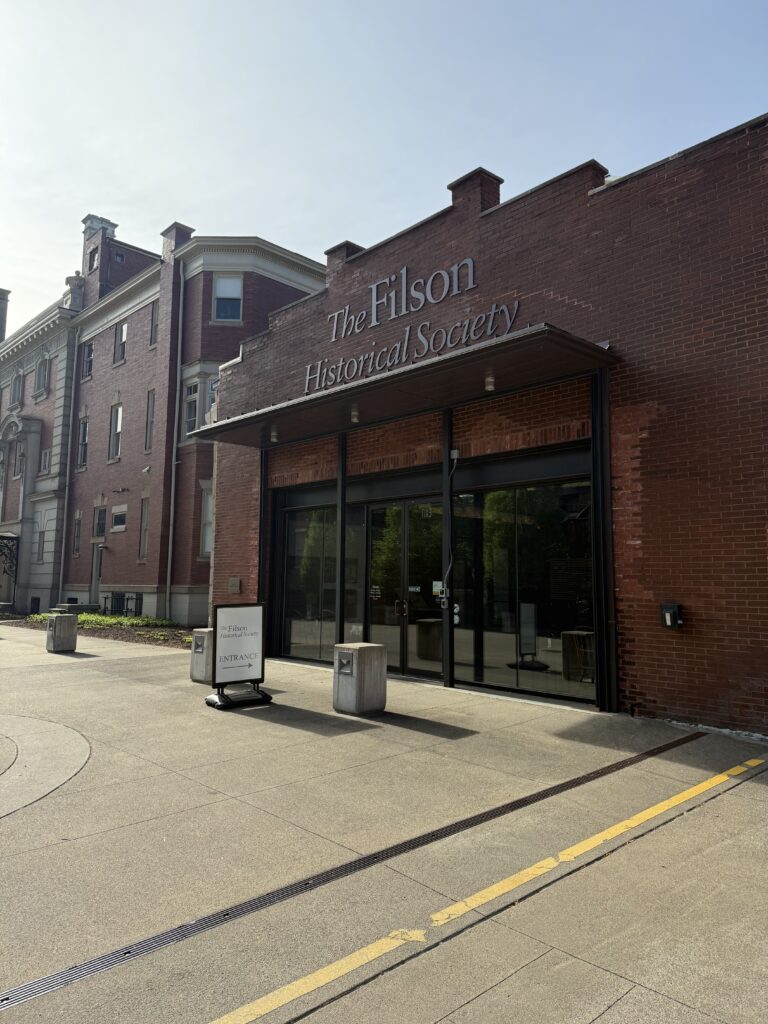 Entrance to The Filson Historical Society, a brick building with large glass doors and a sign out front. The sidewalk and part of the street are visible on a sunny day.