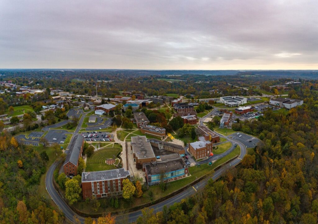Aerial view of a large college campus surrounded by trees and greenery in autumn, with multiple academic buildings, parking lots, and roads under a cloudy sky.