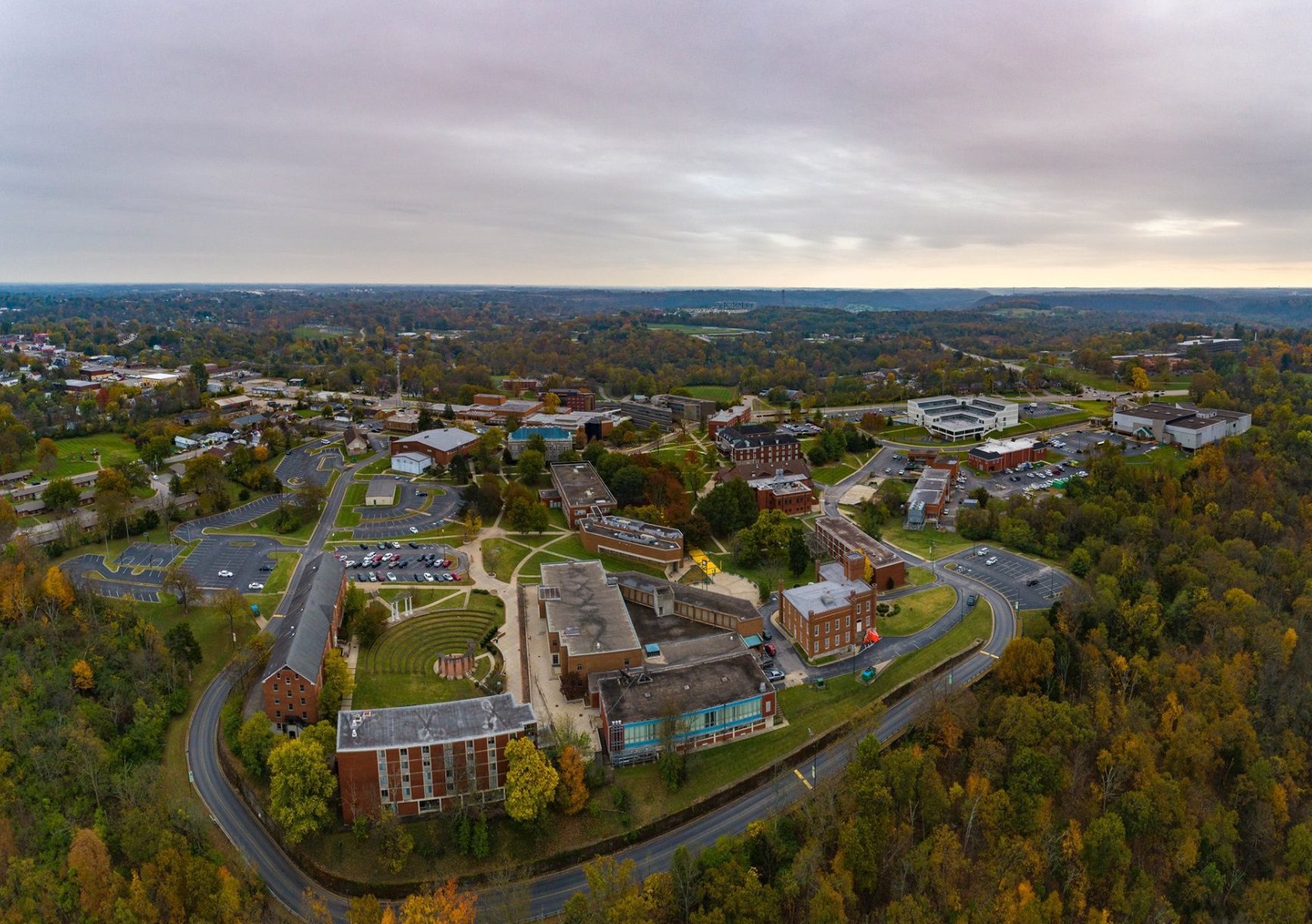 Aerial view of a large college campus surrounded by trees and greenery in autumn, with multiple academic buildings, parking lots, and roads under a cloudy sky.