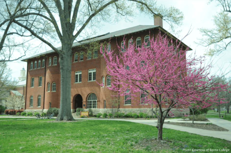 A large brick building with arched windows stands behind a blooming tree with bright pink flowers in a grassy area, surrounded by tall trees and paths on a college campus.