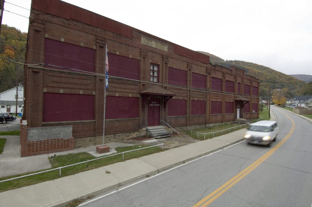 A large, abandoned brick building with boarded-up windows sits beside a street with a car driving by. The building appears weathered, and the area is quiet with trees and hills in the background.