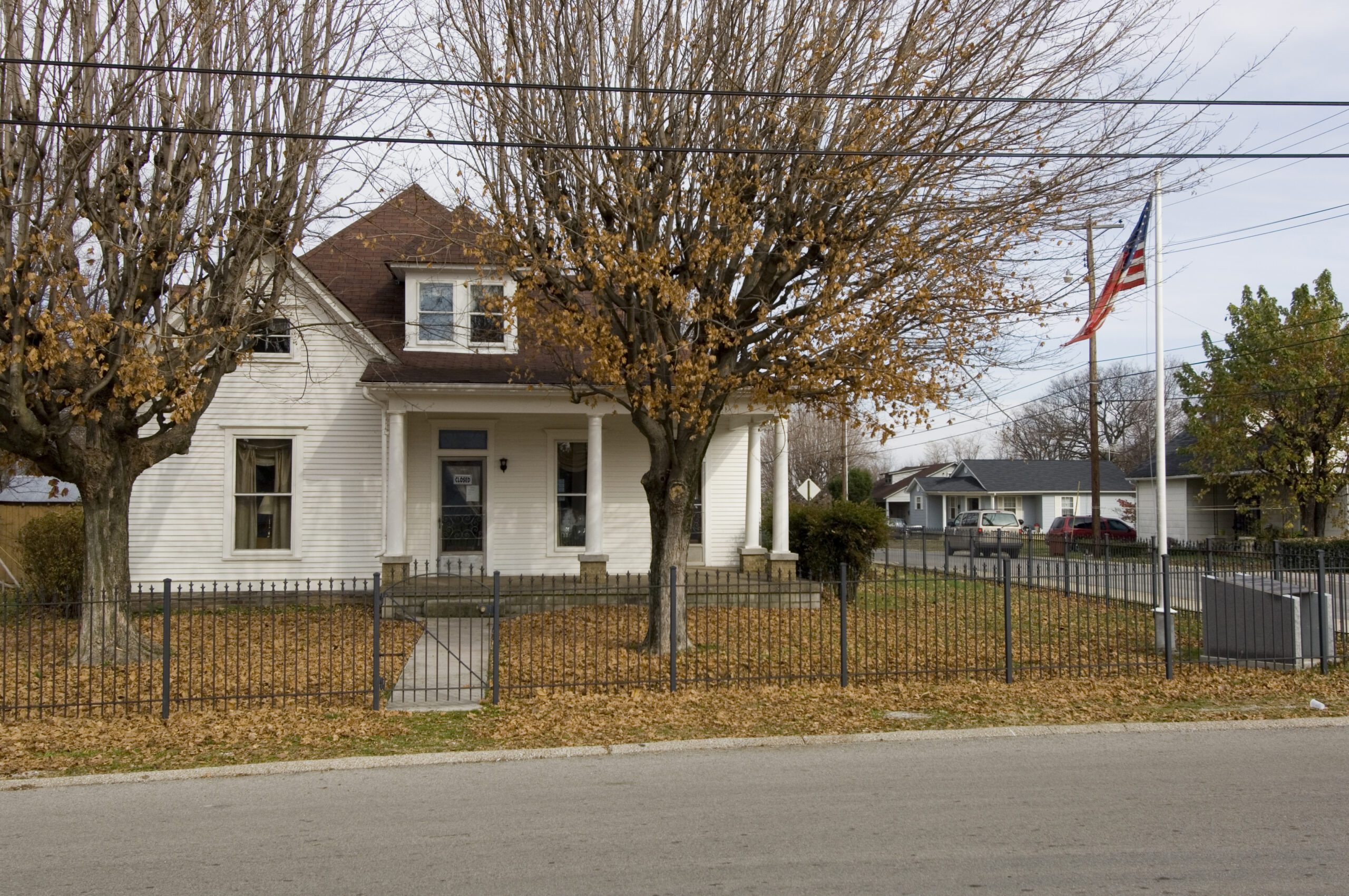 A white two-story house with leafless trees in front, surrounded by a metal fence. Fallen leaves cover the yard, and an American flag is seen on a pole by the driveway. The sky is overcast.