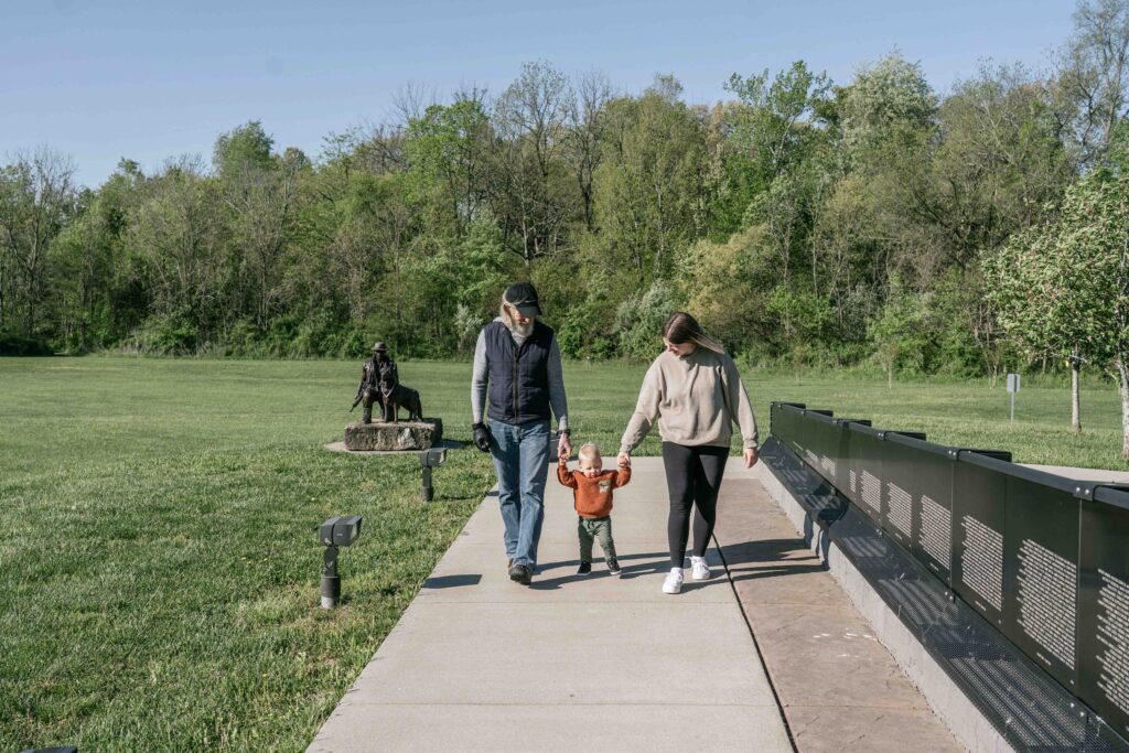 A family of three walks along a pathway in a park. The father holds the childs hand on the left, and the mother holds the childs hand on the right. Behind them is a green field with trees and a statue. It’s a sunny day.