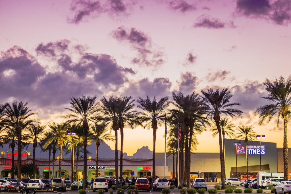 Palm trees and parked cars line a shopping center at sunset, with stores including a fitness gym and Tony & Guy visible under a colorful purple and orange sky with scattered clouds.