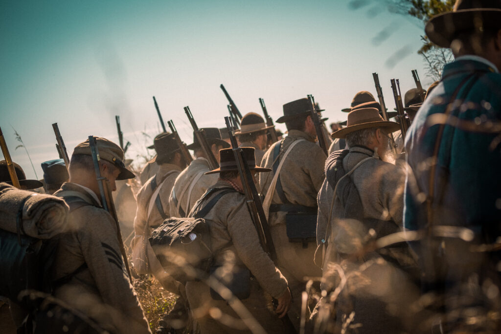 A group of reenactors in period military uniforms march with rifles over their shoulders. They wear wide-brimmed hats, and the scene has a vintage atmosphere with tall grasses in the foreground and a clear sky.