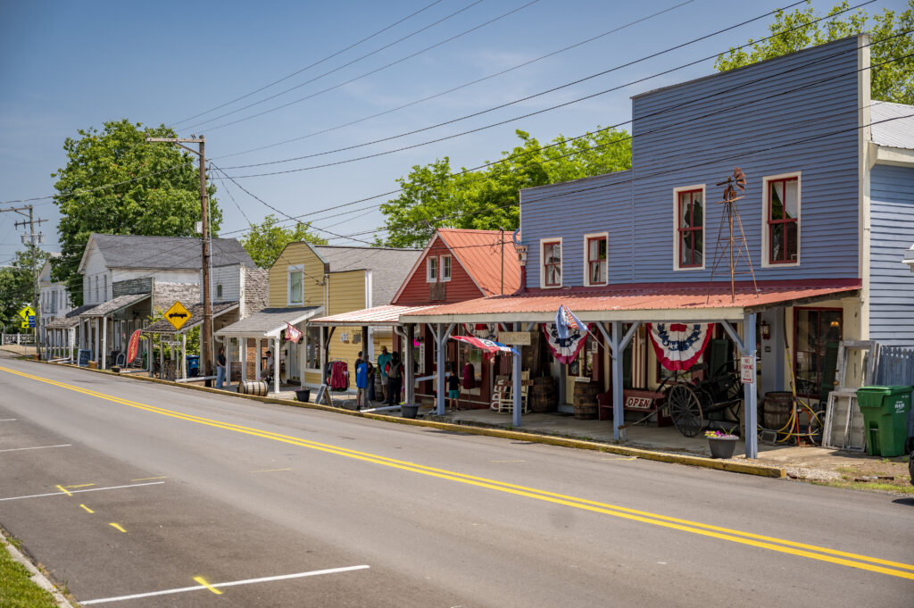 A small-town street with colorful historic buildings, decorated with patriotic bunting. The sunny scene includes a wide road, wooden sidewalks, and people browsing shops under a red awning.