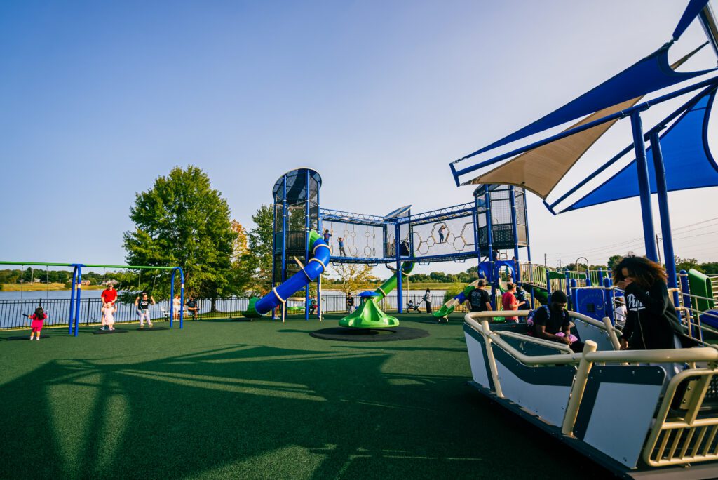 Children playing on a colorful playground featuring slides, swings, and climbing structures under a clear blue sky. A shaded seating area is visible, with trees and a lake in the background.