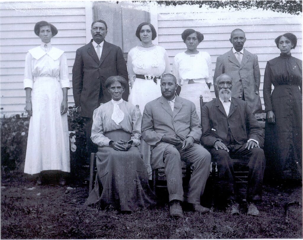 A vintage black-and-white photo shows eight adults posing outside in formal attire. Three women and two men are seated in the front, with three women and two men standing behind them in front of a wooden building.