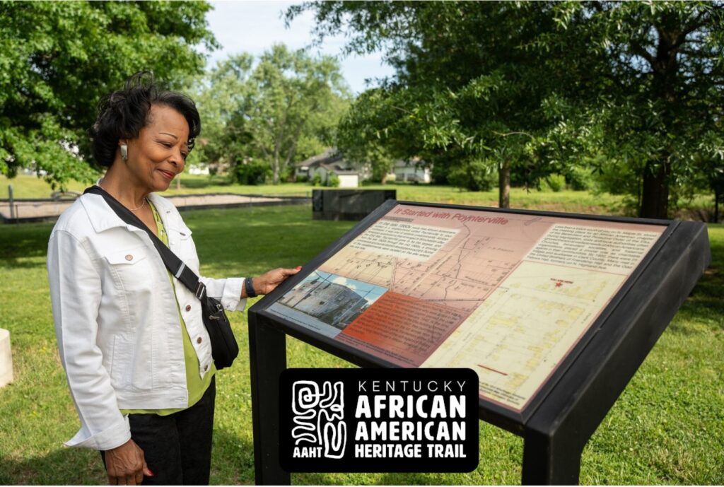 A woman stands outdoors reading an informational sign at a historical site, with trees and grass in the background. The sign features maps and text. A Kentucky African American Heritage Trail logo is displayed in the foreground.