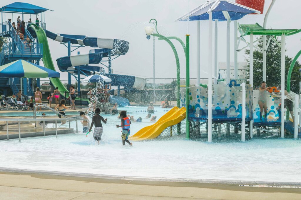 Children play in a water park with slides and splash features. A large blue and green slide towers in the background, and adults and children are enjoying the water attractions on a sunny day.