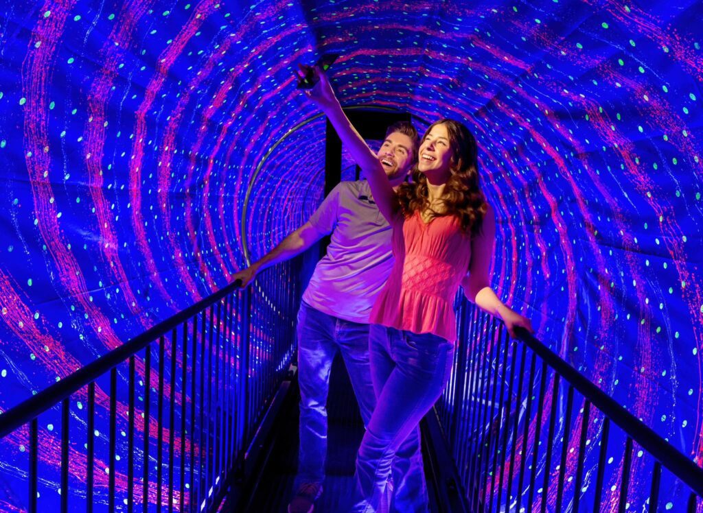 A smiling couple stands on a bridge inside a tunnel with swirling blue and purple lights, pointing and looking excitedly at the vibrant display around them.