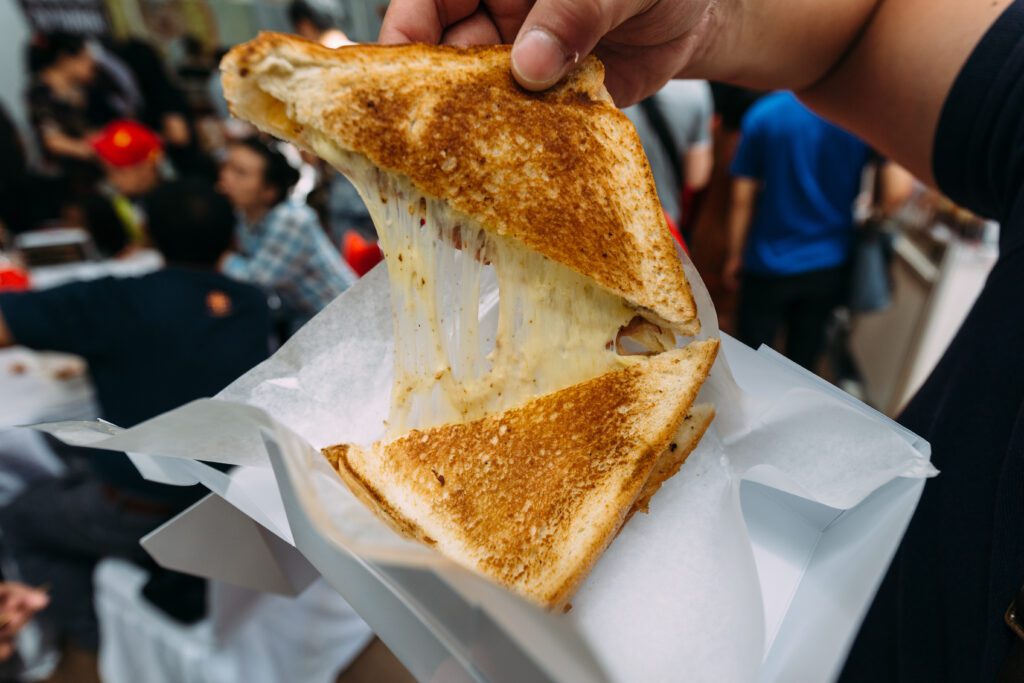 A person holds a grilled cheese sandwich with golden-brown toast, pulling it apart to reveal gooey, melted cheese. The sandwich is served on wax paper, with a blurred background of people sitting and moving around.