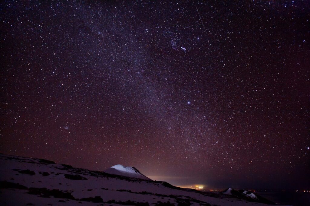 A clear night sky filled with stars and the Milky Way, above a snow-covered mountain landscape with distant lights glowing on the horizon.