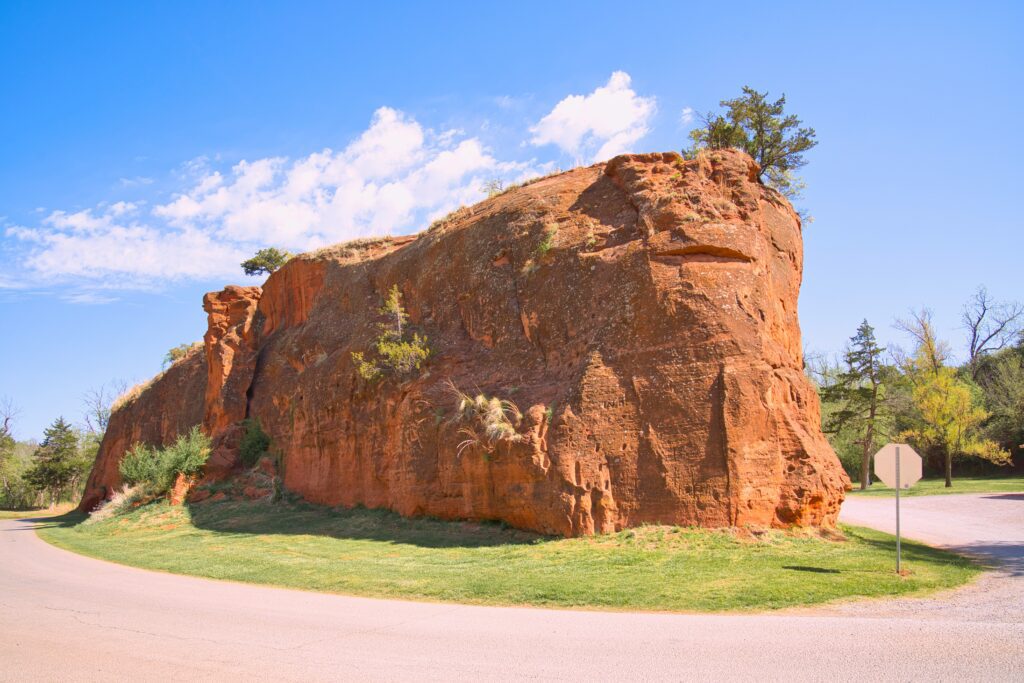 Large red sandstone rock formation sits beside a curved road, with green grass and sparse vegetation around it, under a bright blue sky with scattered clouds. A stop sign is visible to the right.