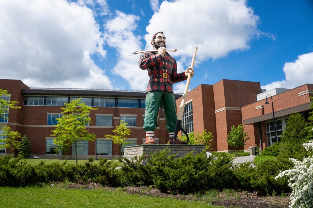 Statue of a bearded lumberjack in a red plaid shirt and green pants, holding an ax and a large pole, standing on a grass-covered pedestal. The backdrop features a brick building and a blue sky with scattered white clouds.
