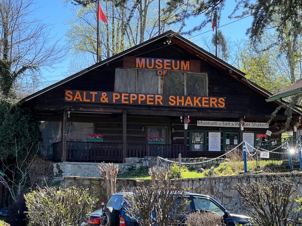 A rustic wooden building with a large orange sign reading Museum of Salt & Pepper Shakers above the entrance, surrounded by trees and bushes on a sunny day.