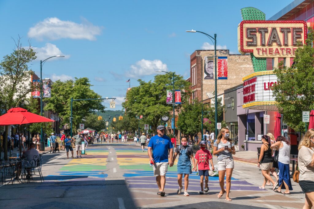A bustling street scene features people walking and enjoying a sunny day. The street is decorated with colorful artwork. In the background, the marquee of the State Theatre is visible. Trees and outdoor seating line the sidewalks.