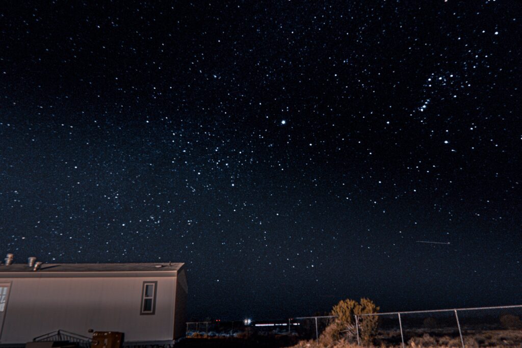 A clear night sky filled with stars above a building and a fence, with a small tree in the foreground. A faint shooting star or meteor is visible near the horizon on the right side of the image.