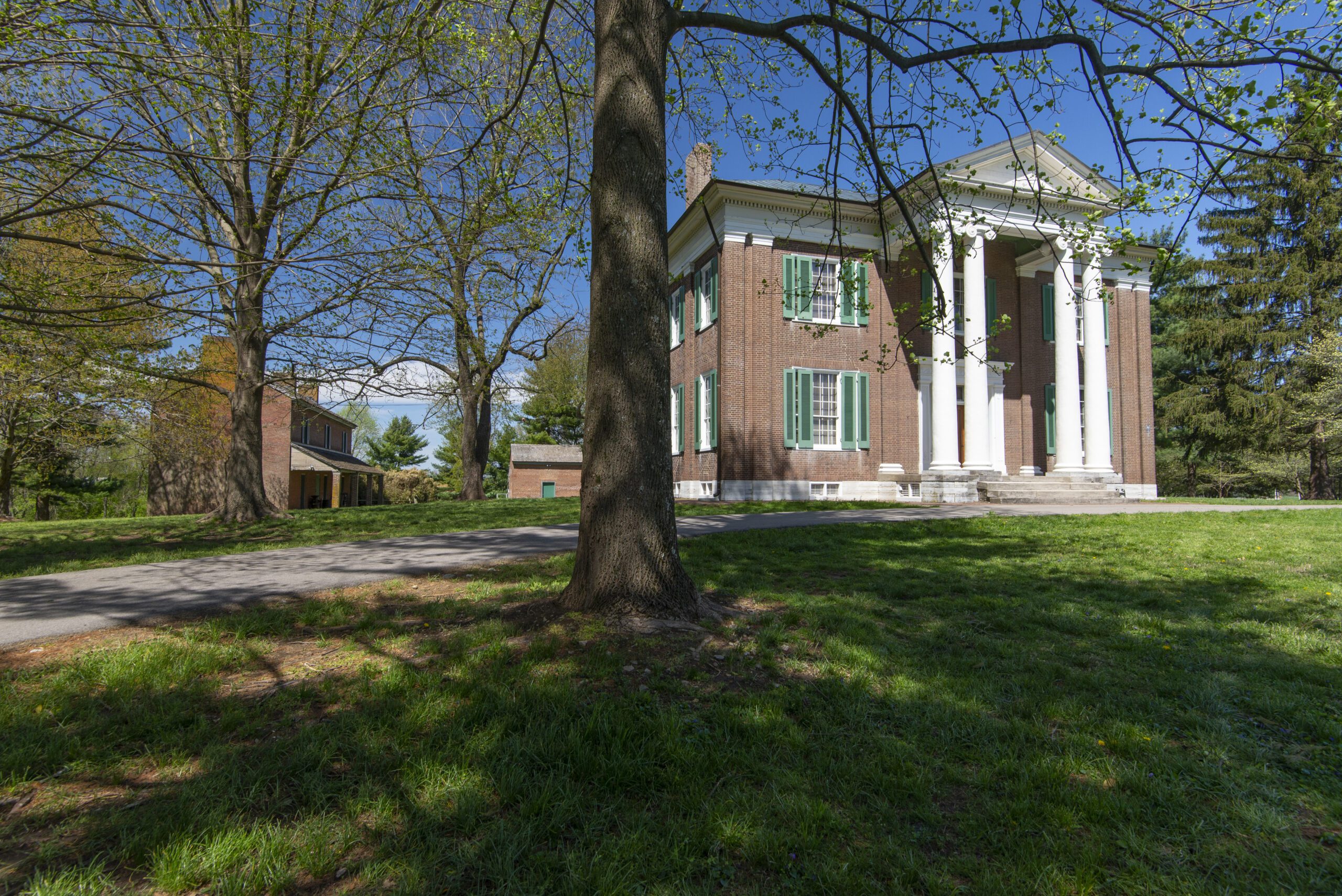 A large red-brick mansion with tall white columns stands in a grassy area, surrounded by trees and a paved path, under a clear blue sky. Another brick building is visible in the background.