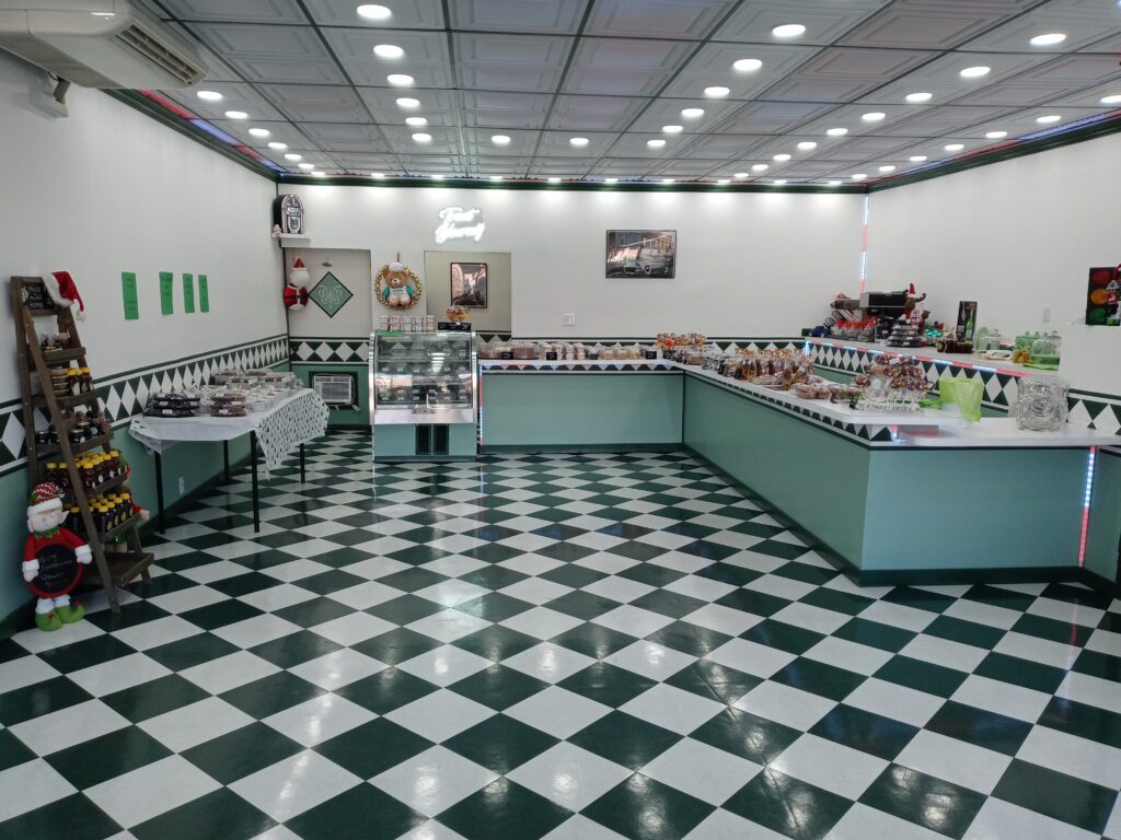 A brightly lit bakery with black and white checkered floor, mint-green counters displaying pastries and desserts, and shelves with bottles. Festive decorations and framed pictures hang on the walls.
