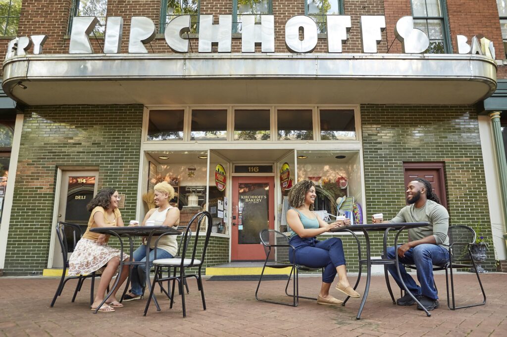 Two pairs of people sit at outdoor tables chatting and smiling in front of a building with a sign reading Kirchhoffs. The scene is relaxed and friendly, with greenery and brick surrounding the sidewalk café.
